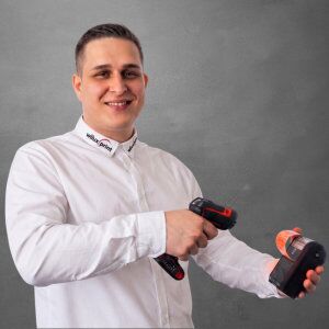 Leotrim Komani, Teil des Teams WILUX PRINT, technischer Verkäufer für AIDC, präsentiert einen Barcode Scanner, professionell gekleidet in einem weissen Hemd vor einem neutralen Hintergrund.